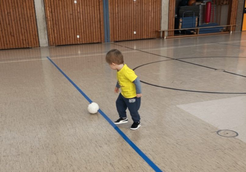 Kleinkind im gelben Trikot kickt einen Ball durch eine Turnhalle
