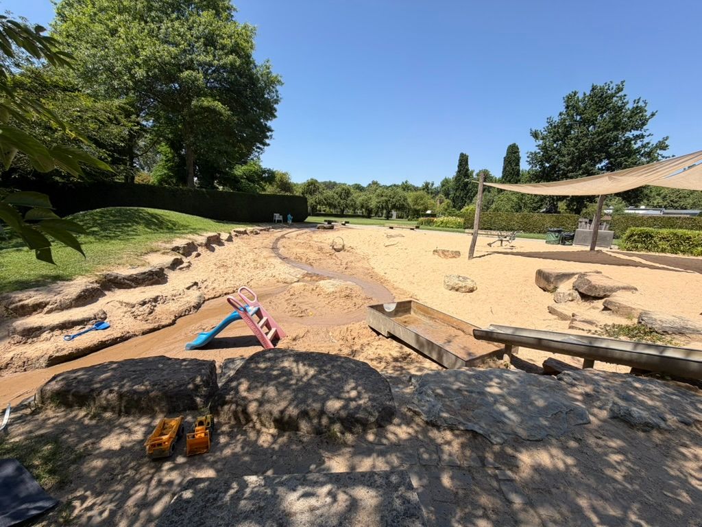 Wasserspielplatz im Park von Schloss Dyck mit Sandfläche, Bachlauf zwischen Steinen und Sonnensegel